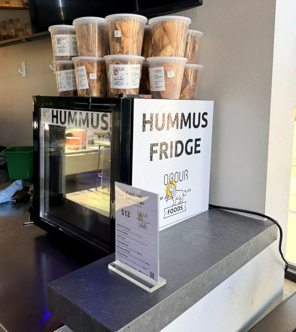 An Obour Hummus Fridge on a bar counter, topped with tubs of pita chips