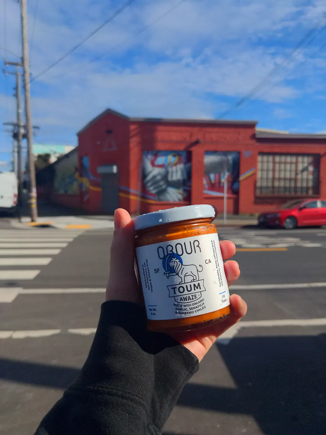 A hand holding a jar of Obour Awaze toum at a crosswalk, with a red brick building behind