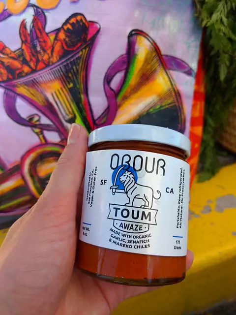 A hand holding a jar of Obour Awaze toum in front of a colorful mural