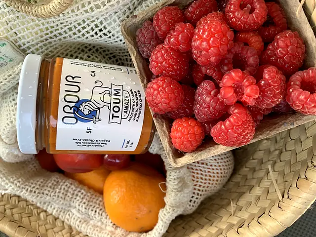 A jar of Obour Awaze toum in a market basket with raspberries and oranges
