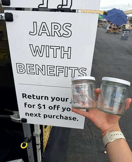 A hand holding two clean empty Obour jars in front of a sign reading Jars with Benefits: return your jar for $1 off your next purchase