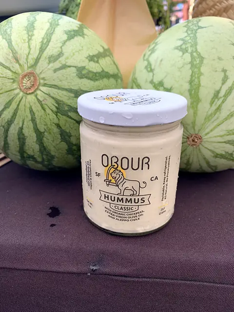 A jar of Obour Classic hummus on a market table between two watermelons