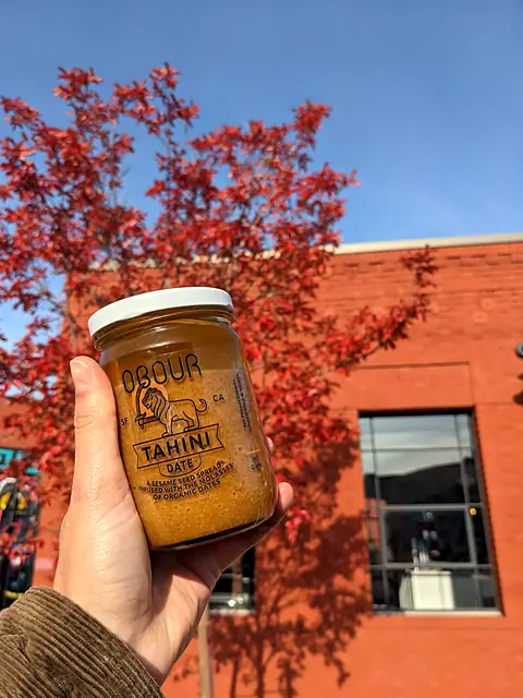 A hand holding a jar of Obour Date tahini in front of a red maple and a brick building