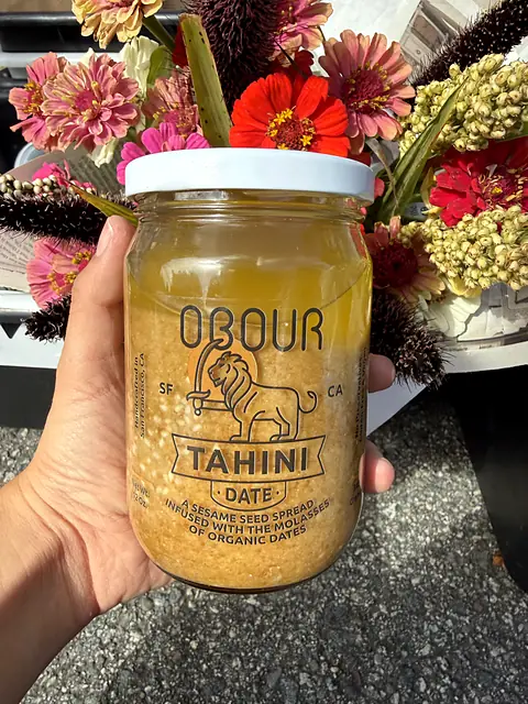 A hand holding a jar of Obour Date tahini in front of a bunch of zinnias