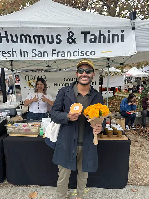 A shopper holding flowers and a tub of Obour hummus in front of the stand