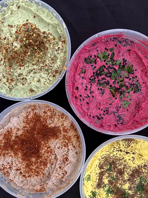 Tubs of Zhoug, Red Beet, Black Bean and Hot Curry hummus from above, garnished with chile, herbs and black sesame