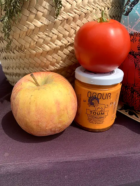 A jar of Obour Gochujang toum with a tomato balanced on top and an apple beside it