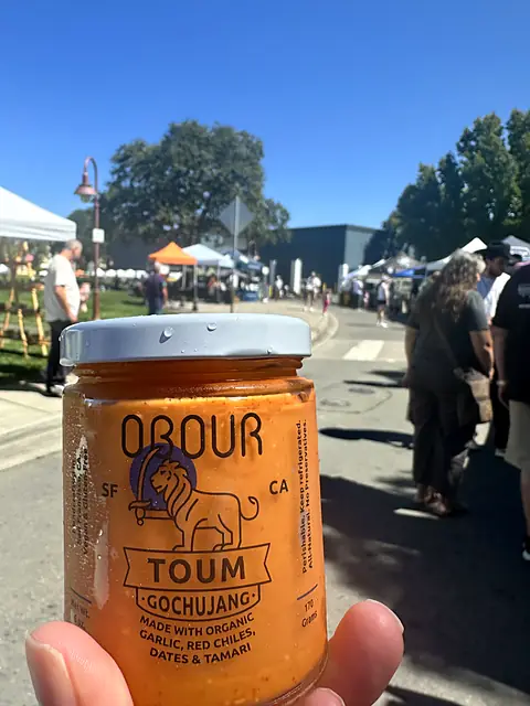 A hand holding a jar of Obour Gochujang toum up at a busy farmers market