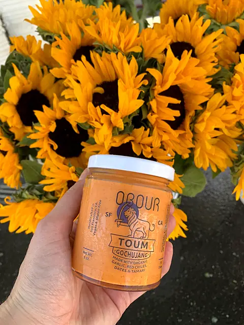 A hand holding a jar of Obour Gochujang toum in front of a bucket of sunflowers