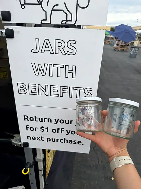 A hand holding two empty Obour glass jars next to the Jars With Benefits jar return sign