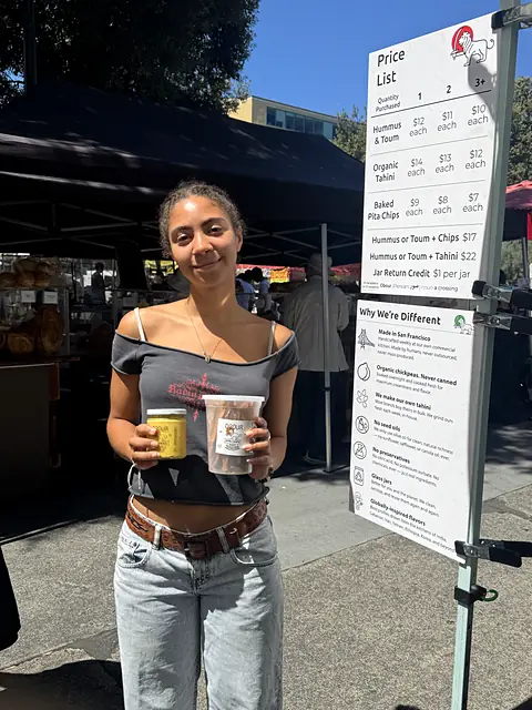 A shopper holding a jar of hummus and a tub of pita chips by the Obour price list