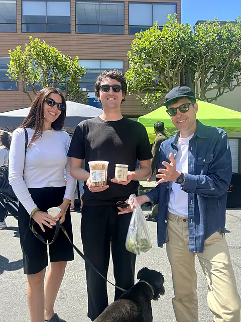 Three friends holding tubs of Obour hummus, with their dog at their feet