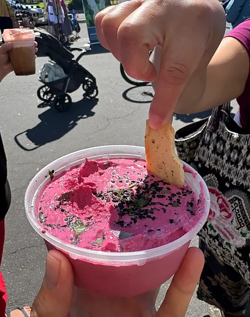 A pita chip dipped into a tub of Red Beet hummus topped with black sesame