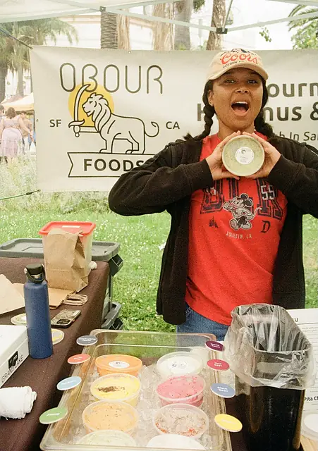 A smiling person holding up a tub of Obour hummus in front of the Obour banner