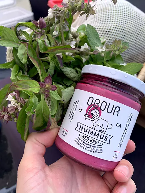 A hand holding a jar of Obour Red Beet hummus in front of fresh basil