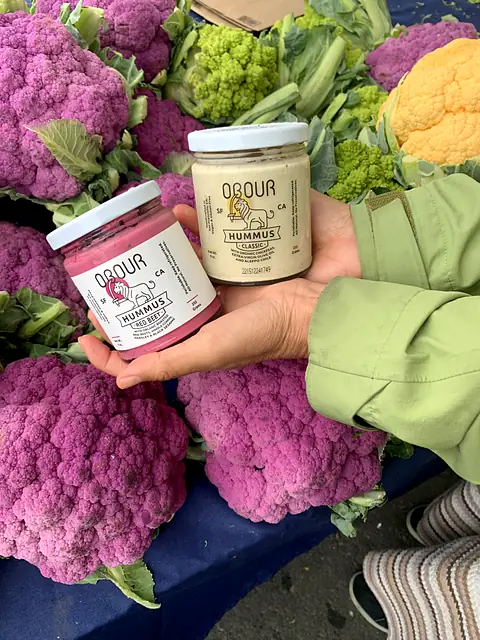 Jars of Obour Red Beet and Classic hummus held up among purple, orange and romanesco cauliflower at a farmers market