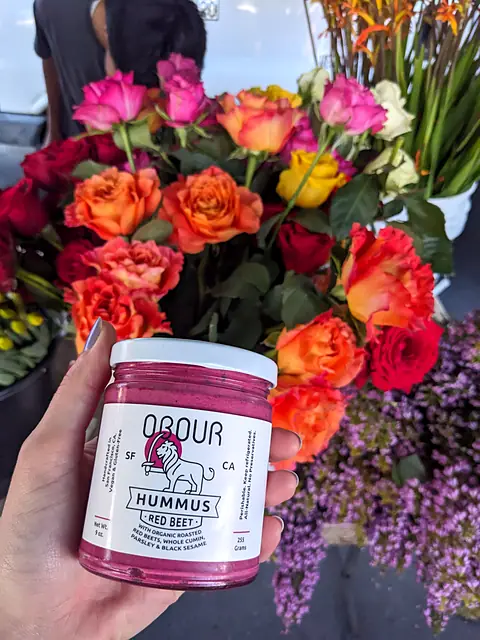 A hand holding a jar of Obour Red Beet hummus in front of a bunch of roses