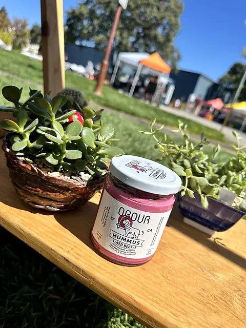A jar of Obour Red Beet hummus on a wooden ledge beside potted succulents at a market