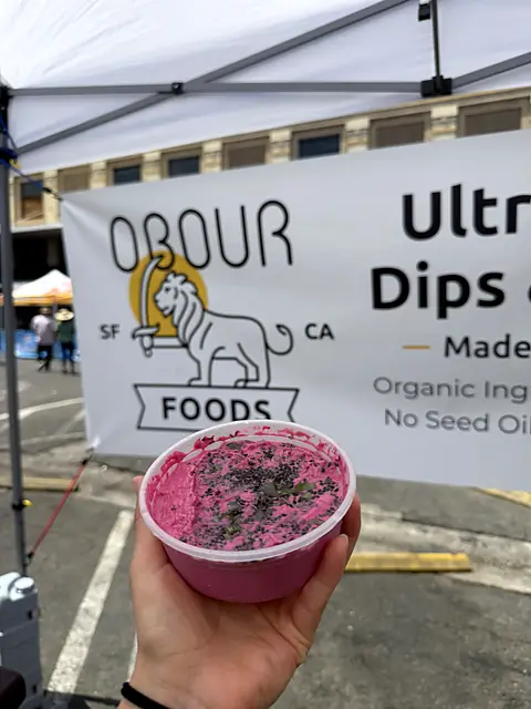 A tub of Obour Red Beet hummus held up in front of the Obour banner