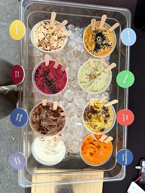 A labeled tray of Obour hummus and toum samples on ice with wooden tasting spoons