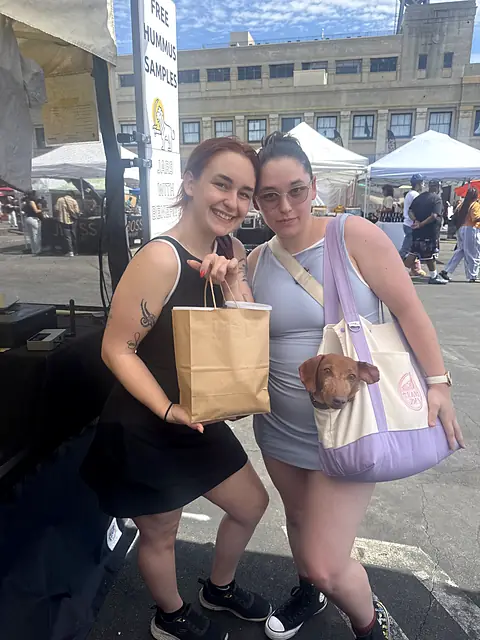 Two friends at the market, one with a shopping bag and one carrying a dog in a tote