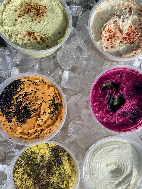 Open tubs of Obour hummus and toum on ice, seen from above, garnished with chile, black sesame and herbs