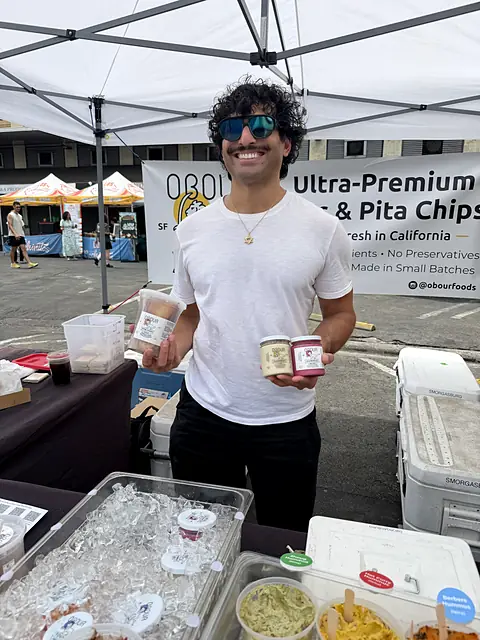 A smiling person holding jars of Obour hummus under the Ultra-Premium Dips & Pita Chips banner