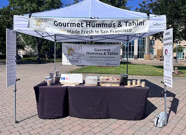 The Obour tent at a farmers market, with a Gourmet Hummus & Tahini banner and a table of jars and pita chips