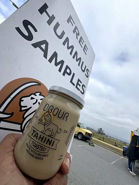 A hand holding a jar of Obour Toasted tahini next to the free hummus samples sign
