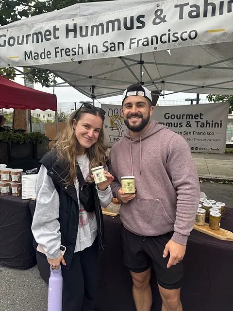 Two shoppers holding jars of Obour hummus in front of the Obour banner