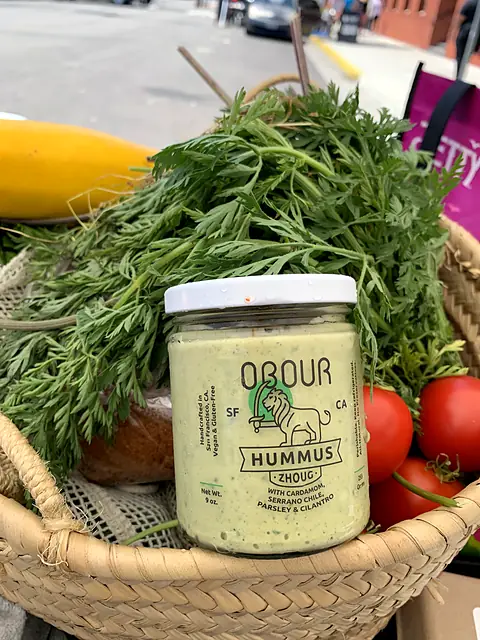 A jar of Obour Zhoug hummus in a basket with carrot tops, squash and tomatoes