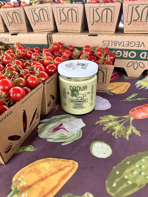A jar of Obour Zhoug hummus next to boxes of cherry tomatoes on a vegetable-print tablecloth