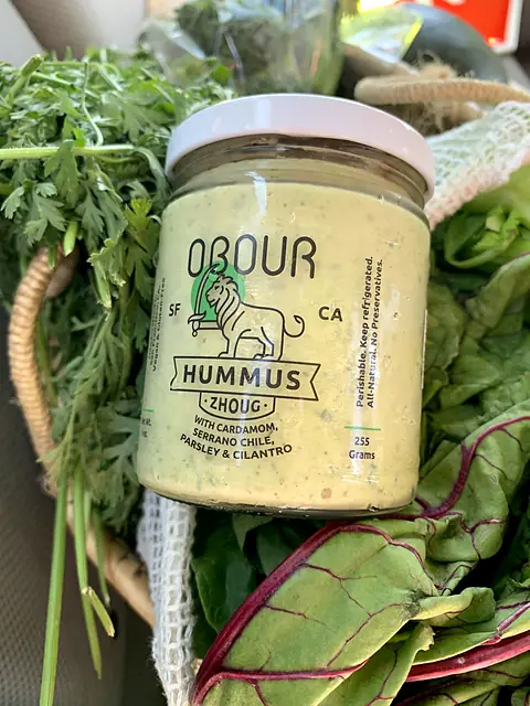 A jar of Obour Zhoug hummus in a basket with carrot tops and chard