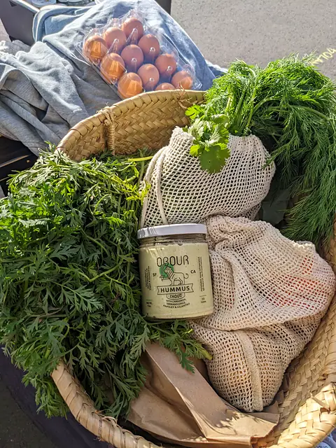 A jar of Obour Zhoug hummus in a woven market basket with carrots, dill and mesh produce bags