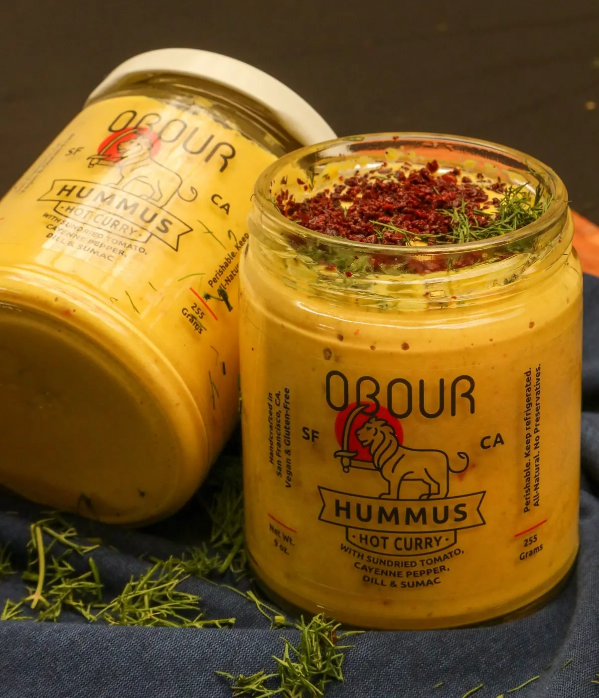 Jars of Obour Hot Curry hummus on a wooden board with fresh herbs