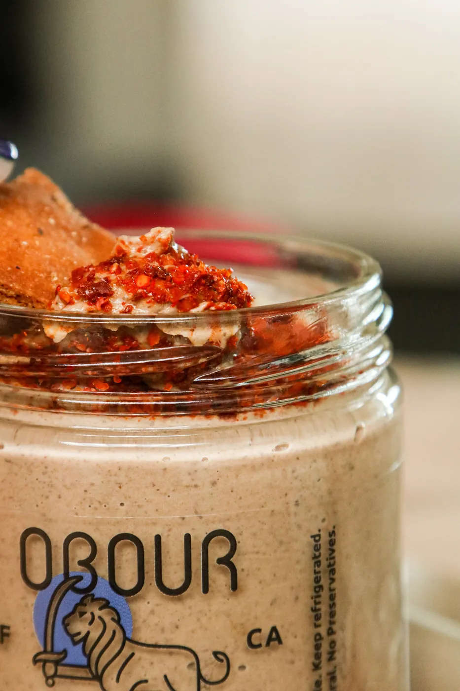 A pita chip dipped into an open jar of Obour Black Bean hummus topped with chile