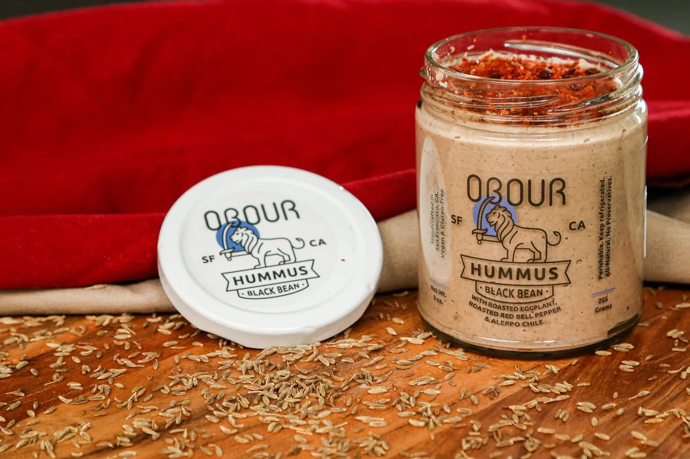 A jar of Obour Black Bean hummus topped with Aleppo chile, its lid propped beside it on a wooden board