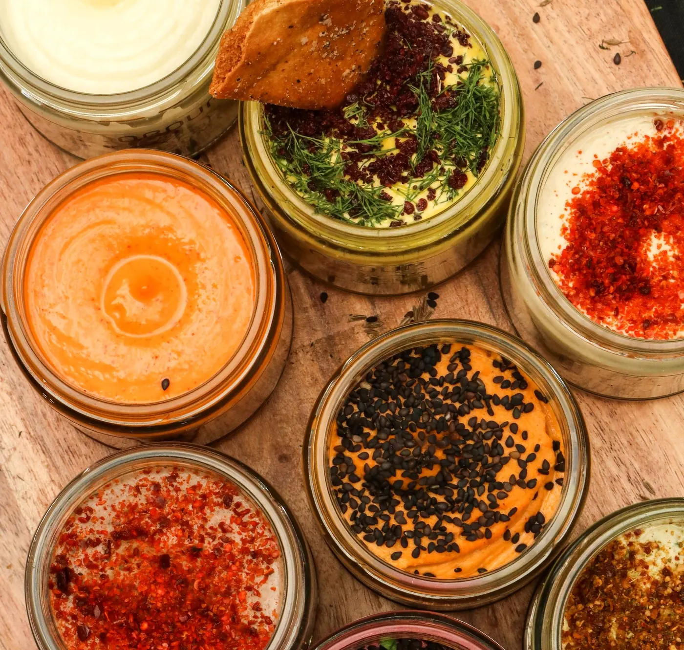 Open jars of Obour hummus and toum on a wooden board, topped with chile, black sesame, beets and dill