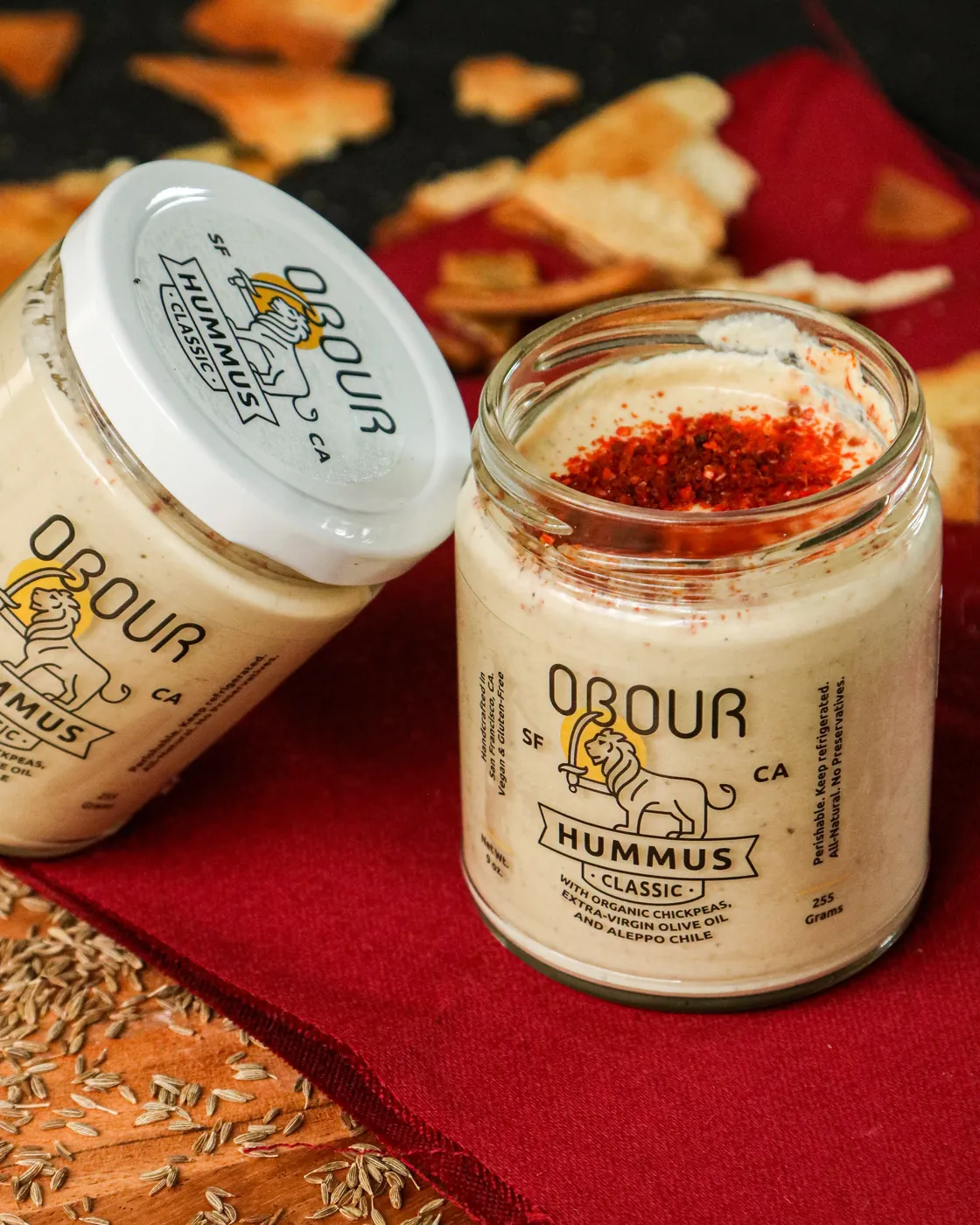 An open jar of Obour Classic hummus dusted with Aleppo chile, next to a closed jar on a red cloth