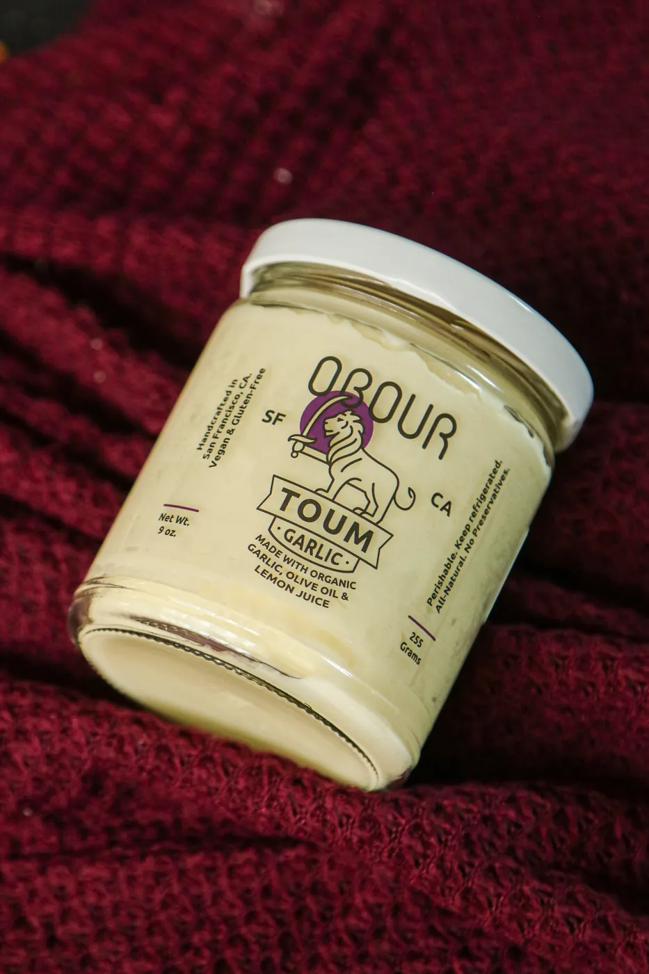 A jar of Obour Garlic toum on a dark red knit cloth, its label reading made with organic garlic, olive oil and lemon juice