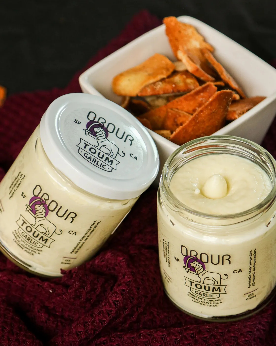 An open jar of Garlic toum next to a closed jar and a bowl of pita chips
