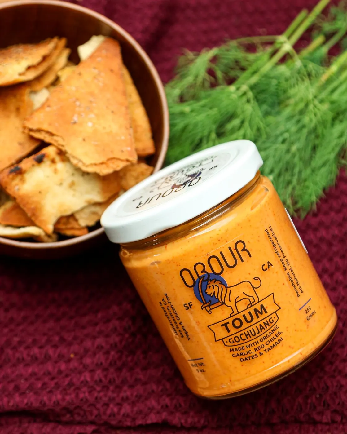 A jar of Gochujang toum next to a bowl of pita chips and fresh dill