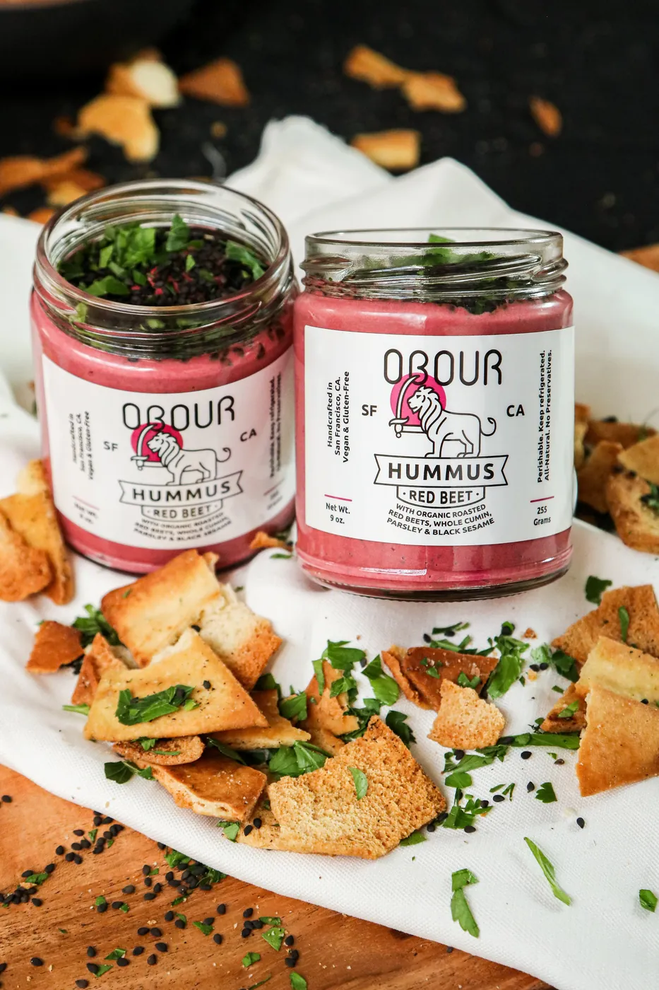 Two jars of Obour Red Beet hummus on a white cloth, surrounded by pita chips, parsley and black sesame