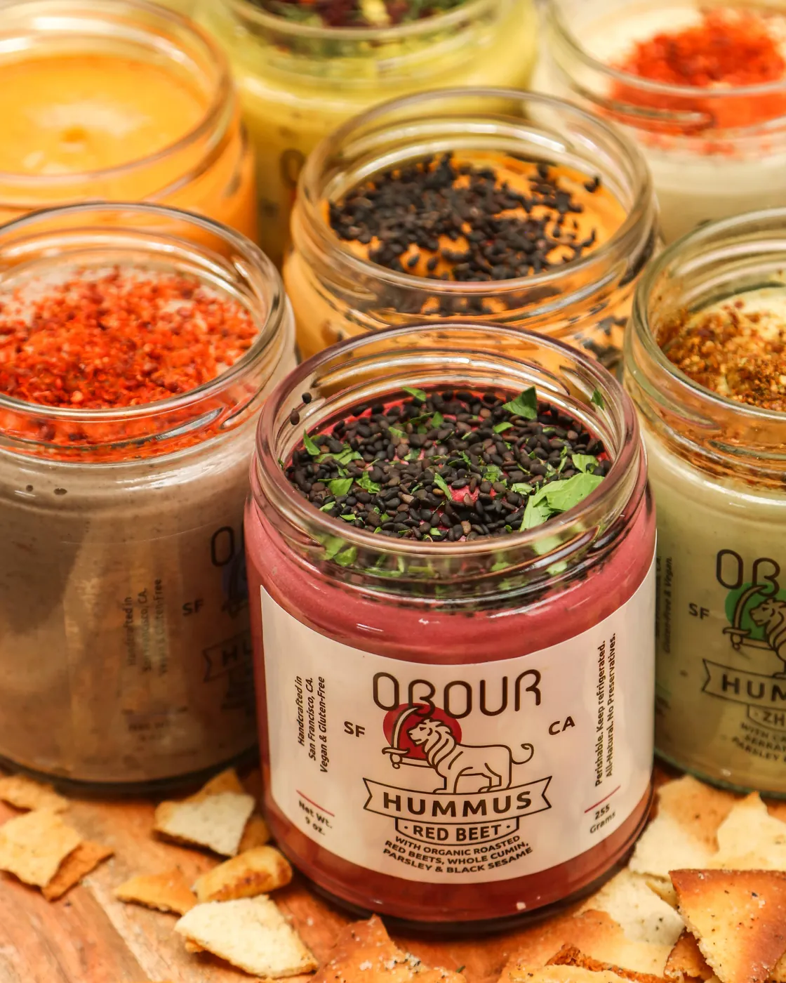 An open jar of Obour Red Beet hummus topped with black sesame and parsley, in front of open jars of the other flavors and pita chips