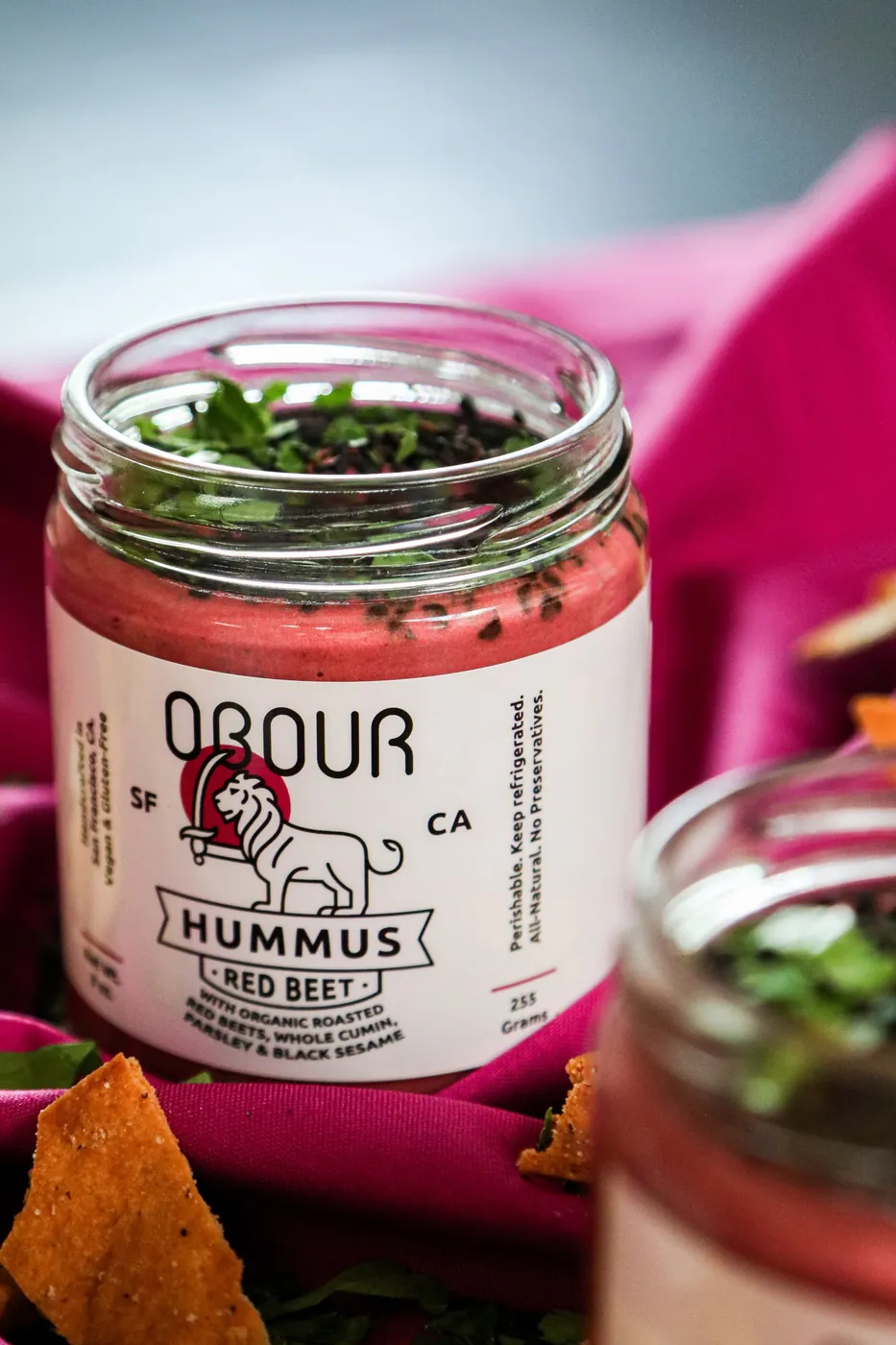 An open jar of Obour Red Beet hummus topped with black sesame and parsley on a pink cloth