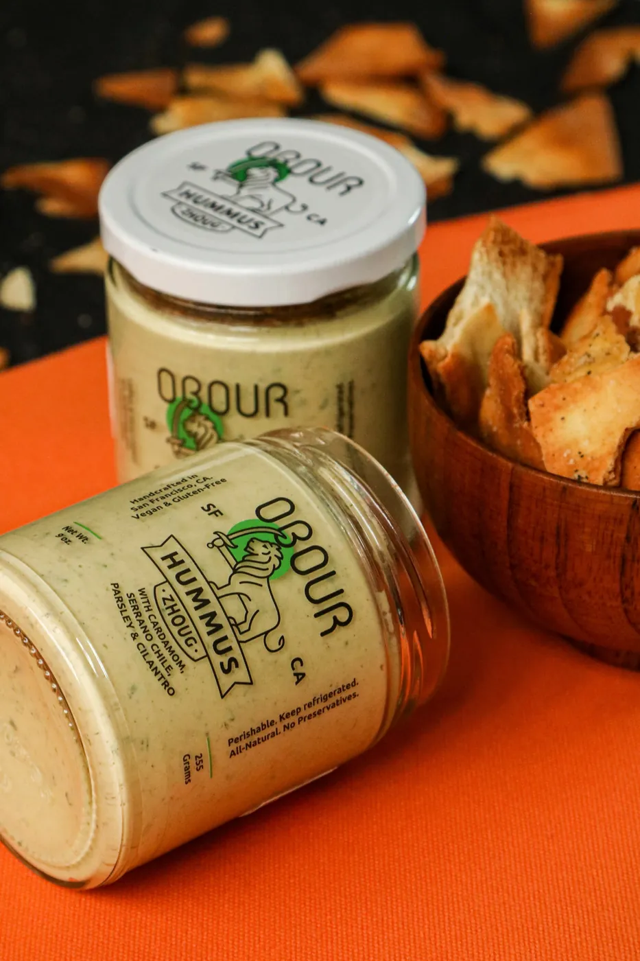 Two jars of Obour Zhoug hummus on an orange cloth next to a wooden bowl of pita chips