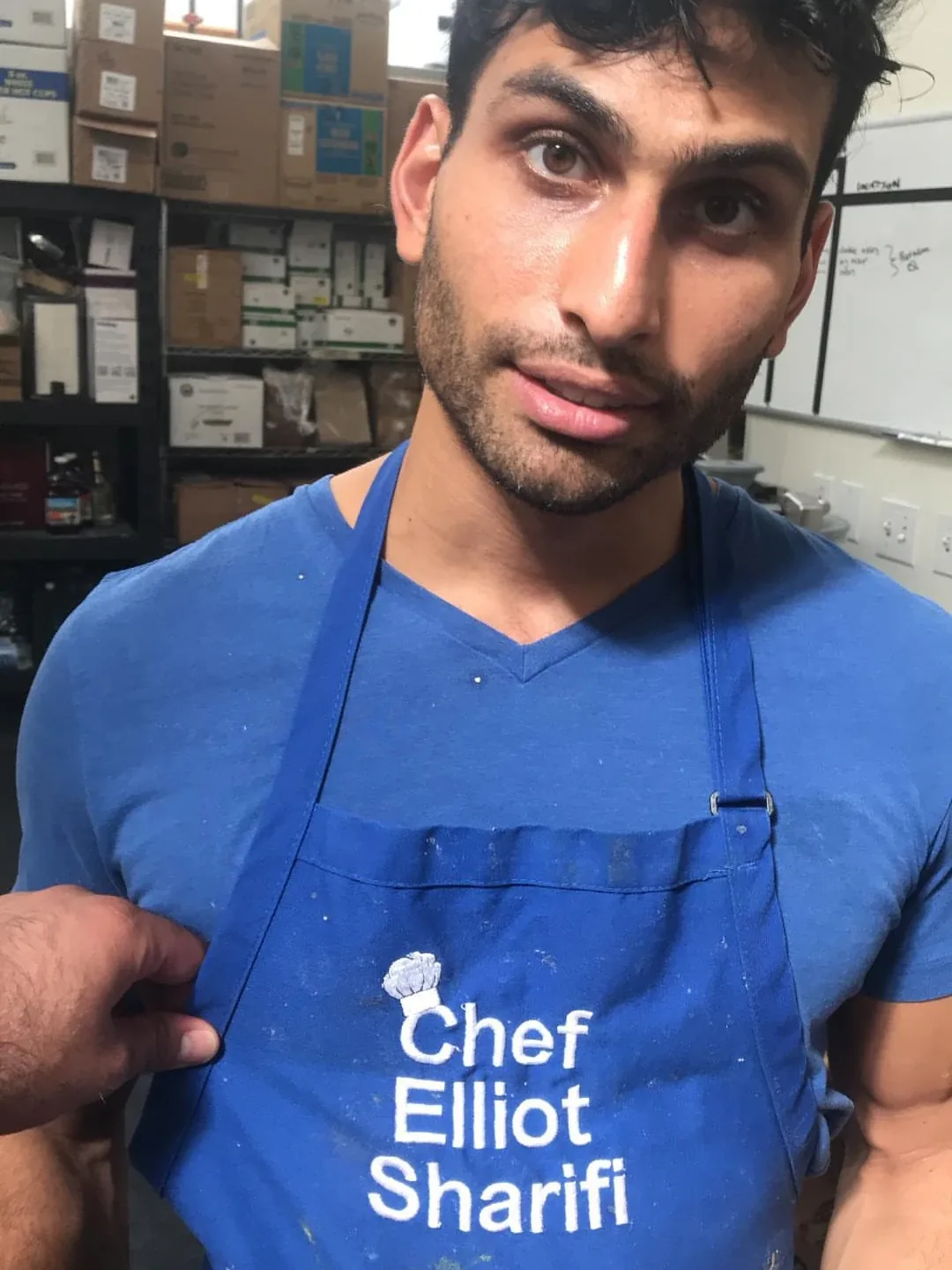 Elliot in a blue apron embroidered “Chef Elliot Sharifi,” in the middle of a long production day around 2018