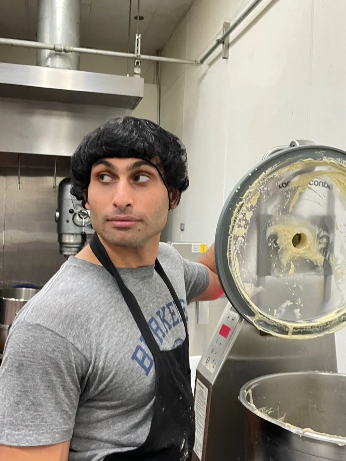 Elliot in a hairnet and apron, opening the lid of a mixer full of hummus in the kitchen, around 2023