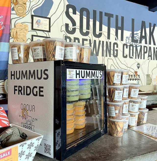 An Obour Hummus Fridge on the bar at South Lake Brewing, stocked with hummus and toum tubs, with pita chips stacked beside it