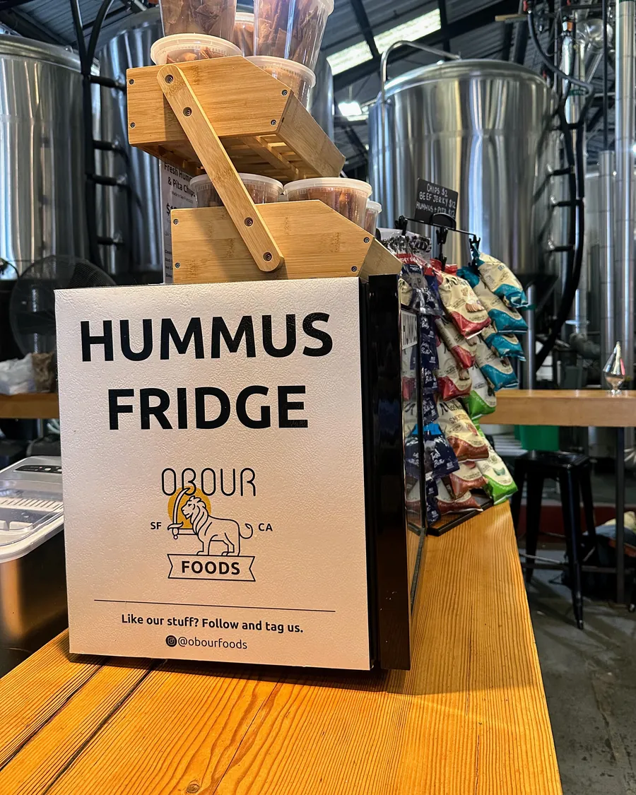 An Obour Hummus Fridge sign on a brewery counter, with brewing tanks behind